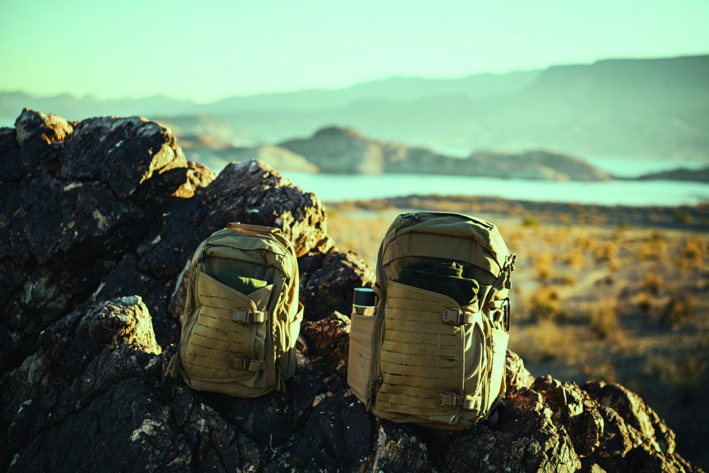 Two tactical backpacks rest on rocky terrain overlooking a lake and mountains, surrounded by dry brush under a clear sky, suggesting field readiness