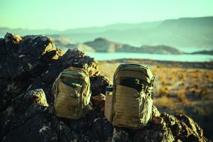 Two tactical backpacks rest on rocky terrain overlooking a lake and mountains, surrounded by dry brush under a clear sky, suggesting field readiness