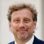 Person in dark blue checkered suit, white shirt, purple striped tie, and small lapel pin on left side, posed against neutral backdrop
