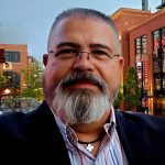 Person wearing a dark blazer over a striped collared shirt with a silver cross necklace, standing outdoors near brick buildings with lit signage and trees.