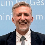 Person in black suit, white shirt, gray tie stands before light blue backdrop with partial white text reading “um für Ges... inium”