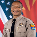 Arizona State Trooper in uniform with badge, nameplate, and shoulder patch stands before U.S. and Arizona flags on a formal backdrop.