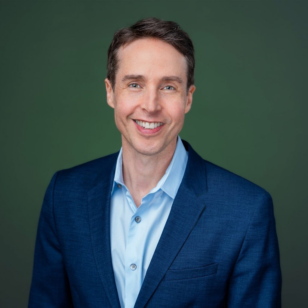 Individual in blue suit jacket and light blue open-collar shirt stands before solid green background