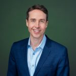 Individual in blue suit jacket and light blue open-collar shirt stands before solid green background