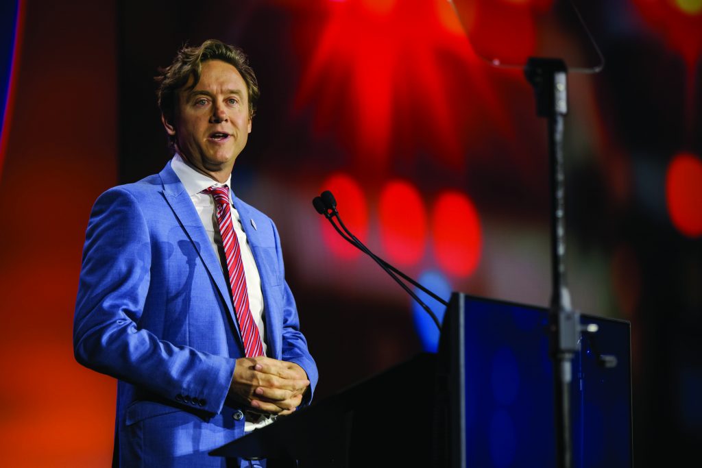 Person in a blue suit and red striped tie stands at a podium with dual microphones on stage, bright red and orange lights creating a vibrant backdrop