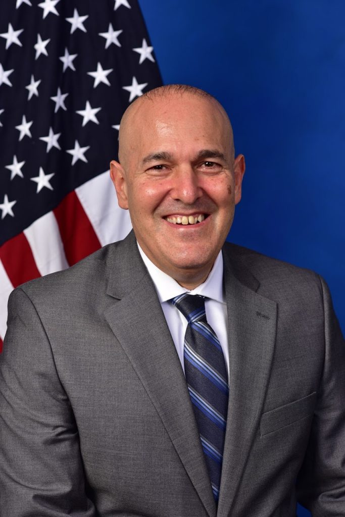 A professional portrait of an individual in a gray suit with a white dress shirt and a blue striped tie. The person is seated against a backdrop featuring the American flag on the left side and a solid blue background on the right. The American flag has visible stars and stripes, adding to the formal and patriotic tone of the image.