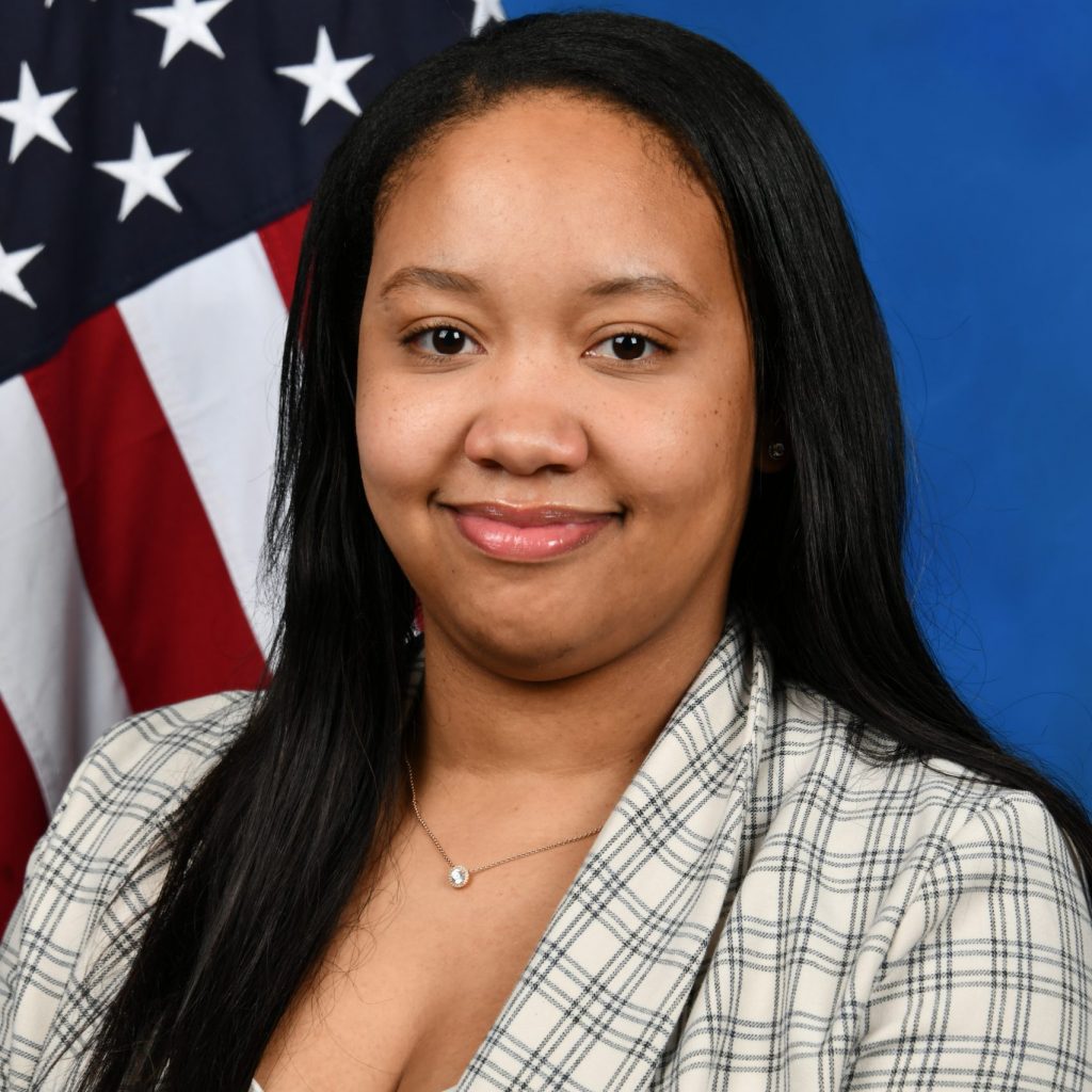 a person with long black hair wearing a plaid blazer over a white top. They are standing in front of an American flag, which is partially visible in the background. The background also includes a blue backdrop. The person is wearing a delicate necklace with a small pendant.