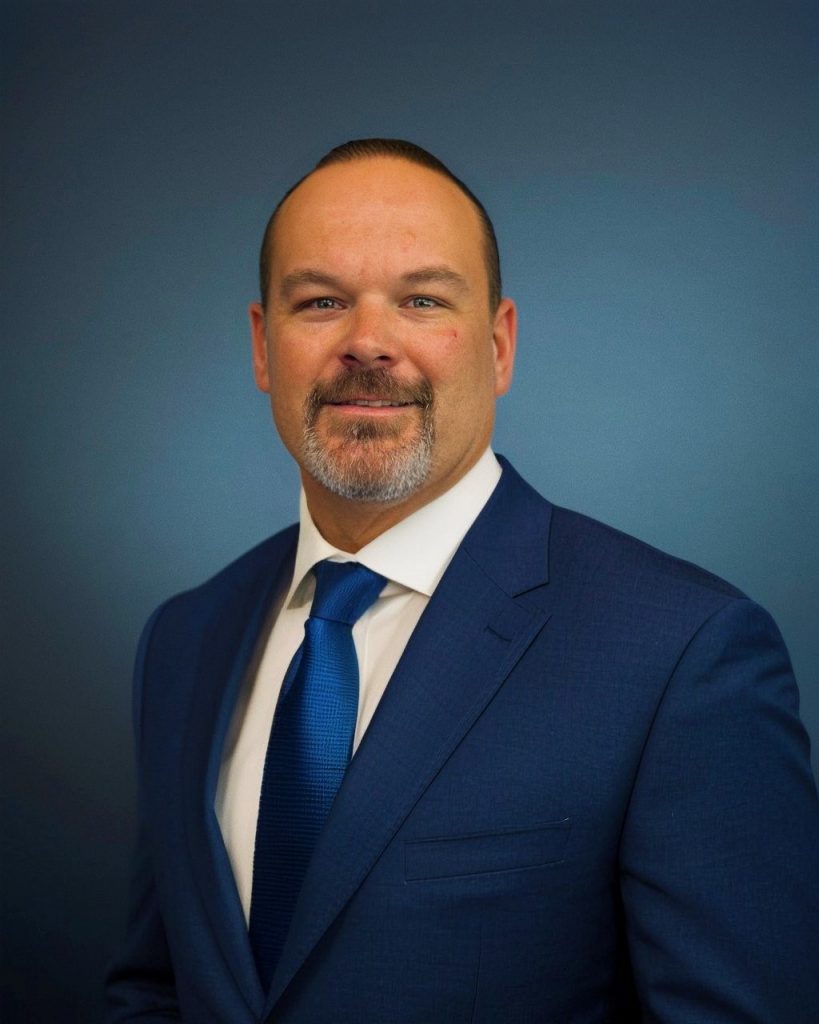 Person wearing a tailored blue suit, white dress shirt, and blue tie posed against a smooth dark blue backdrop in a formal portrait setting.