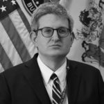Individual in suit and tie stands before American flag and a second flag with intricate design