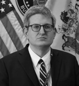 Individual in suit and tie stands before American flag and a second flag with intricate design