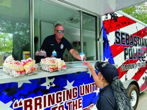 A police officer is serving a woman from a food truck labeled "Sebastian Police Community Policing." The truck has an American flag design with red, white, and blue colors. The officer, wearing a black uniform with a badge and name tag, is handing over a blue snow cone to the woman. The woman, dressed in black with long braided hair and wearing a cap that says "JACK," is reaching out to take the snow cone. On the counter of the food truck are several baskets filled with popcorn. The text on the side of the truck reads "BRINGING UNITY TO THE COMMUNITY." Trees are visible in the background through the windows of the food truck. Both the officer's and the woman's faces are visible, showing friendly expressions.