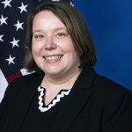 a person in front of an American flag. The person is wearing a black blazer over a black top with a white scalloped neckline. The background is blue, and the American flag is partially visible on the left side of the image.