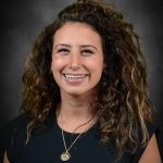 Person with long curly hair wearing a black top and gold necklace stands before a dark textured backdrop in a formal portrait‑style photograph