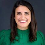 Person wearing a bright green blouse with pleated shoulders and lace detailing, standing against a dark gray background in a professional portrait style.