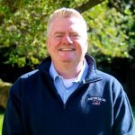 Individual in navy "Maritime College" zip-up jacket over light blue collared shirt stands outdoors in front of dense green foliage