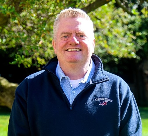 Individual in navy "Maritime College" zip-up jacket over light blue collared shirt stands outdoors in front of dense green foliage