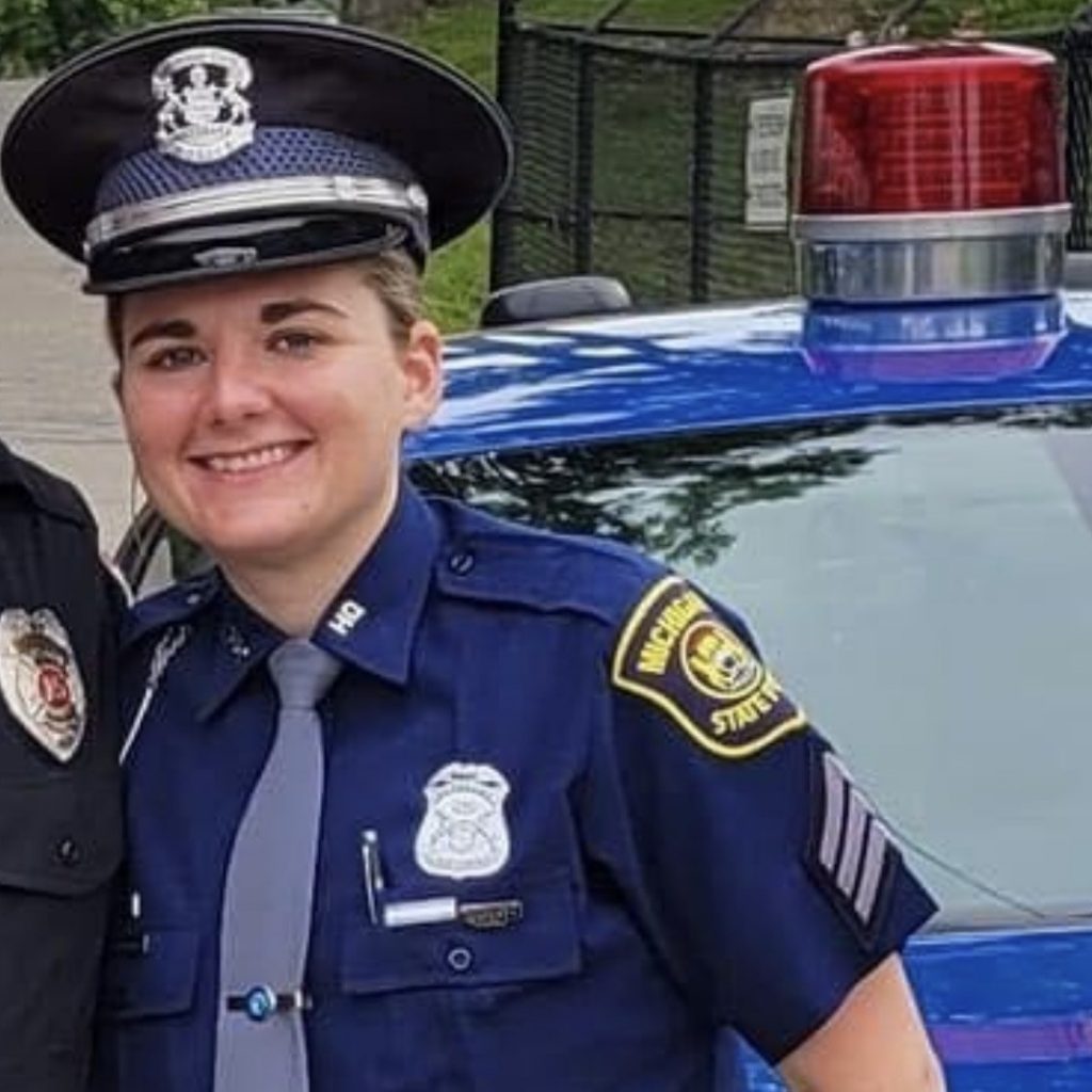 Michigan State Police officer in uniform stands beside patrol car with red light; badge, shoulder patch, and trees in background highlight official setting