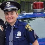 Michigan State Police officer in uniform stands beside patrol car with red light; badge, shoulder patch, and trees in background highlight official setting