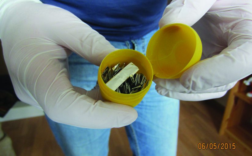 Gloved hands hold open yellow plastic container dated 06/05/2015, containing small metallic objects and a white strip, suggesting evidence handling