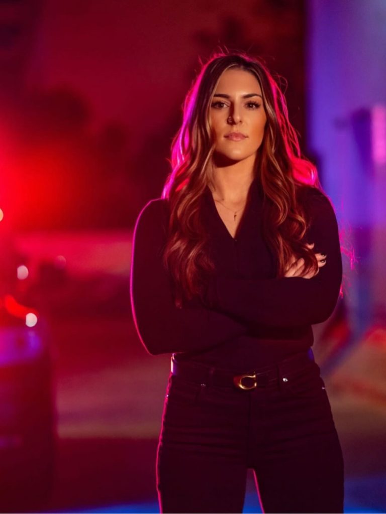 Person standing outdoors at night with arms crossed, wearing dark long-sleeve top and pants; red and blue emergency lights illuminate the background