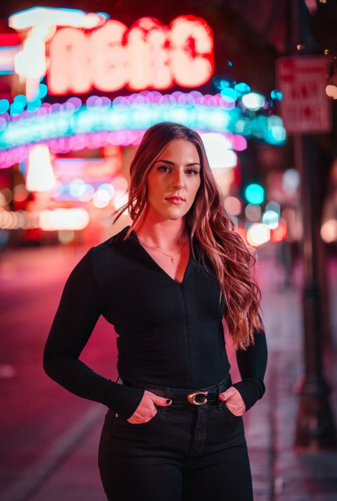 Person standing on a city sidewalk at night wearing a black long-sleeve top, black pants, and patterned belt; neon lights and street signs glow in background