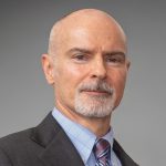 Person wearing a dark suit jacket over a light blue checkered dress shirt with a patterned tie, posed against a plain gray background