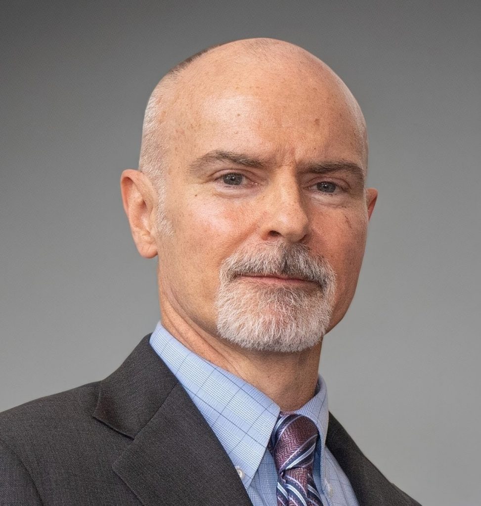 Person wearing a dark suit jacket over a light blue checkered dress shirt with a patterned tie, posed against a plain gray background