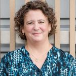 Person wearing a blue patterned blouse with a gold necklace featuring a small pendant, standing in front of vertical wooden slats and gray background