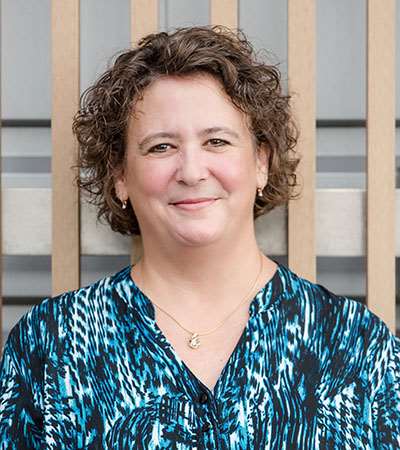 Person wearing a blue patterned blouse with a gold necklace featuring a small pendant, standing in front of vertical wooden slats and gray background