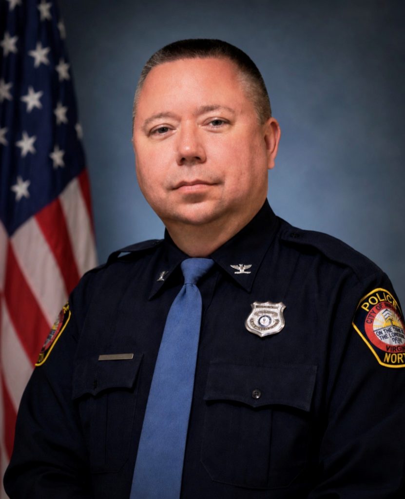 Uniformed sheriff’s official wears a white dress shirt with brown shoulder panels, tie, gold star badge, collar stars, U.S. flag patch, and service ribbon, posed before a U.S. flag and studio backdrop