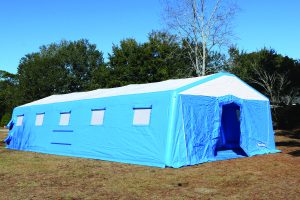 Large blue-and-white inflatable shelter tent set up on open ground, with multiple covered windows and an open entrance, surrounded by trees under a clear sky