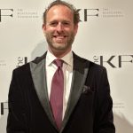 Person wearing a black velvet tuxedo jacket with satin lapels, white dress shirt, burgundy tie, and pocket square, standing before Kosciuszko Foundation backdrop.