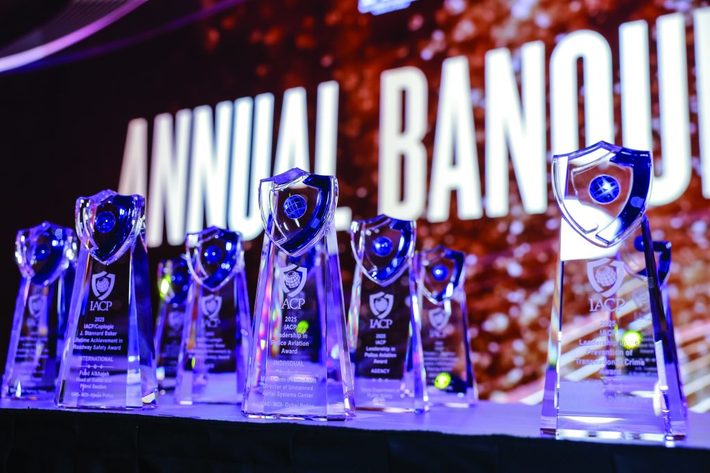 Row of clear glass awards with shield emblems and engraved text displayed on a black table; large illuminated “ANNUAL BANQUET” sign in background.