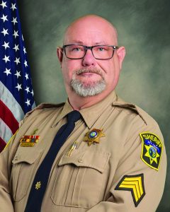 Sheriff in uniform stands before American flag; badge, shoulder patch, rank insignia, pins, and ribbons clearly visible on shirt.