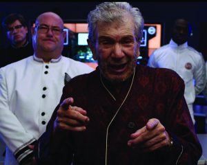 Person in patterned robe points forward, flanked by three in white uniforms; control room with multiple screens in background