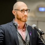 Individual in dark blazer, pink shirt, gray zip-up sweater speaks at podium with mic; purple awareness ribbon pinned to blazer; background not visible.