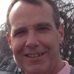 Person wearing a light gray patterned suit jacket over a pink dress shirt with a dark patterned tie, standing outdoors with trees and power lines in background