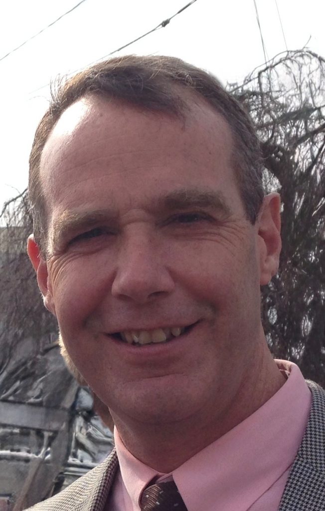 Person wearing a light gray patterned suit jacket over a pink dress shirt with a dark patterned tie, standing outdoors with trees and power lines in background