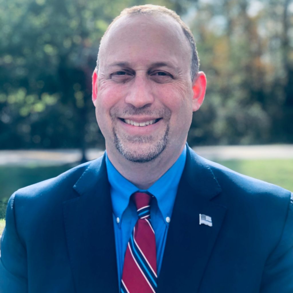 An individual in a dark suit jacket, blue dress shirt, striped red tie, and flag lapel pin stands outdoors with trees, grass, and a sidewalk blurred behind