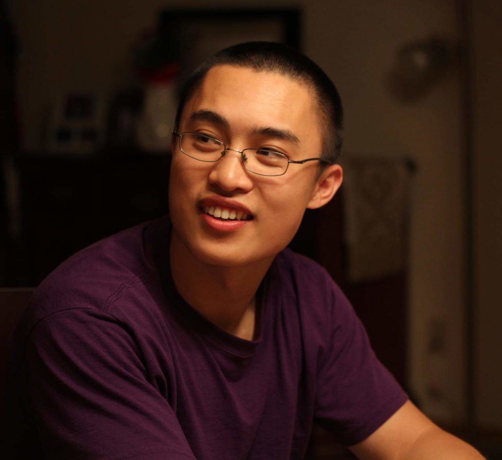 Person seated indoors wearing a plain purple T-shirt; background shows dimly lit room with furniture and a patterned towel hanging on a rack