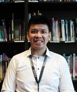 Person in a white collared shirt with a patterned lanyard sits in front of tall bookshelves filled with assorted books in an office or library setting.