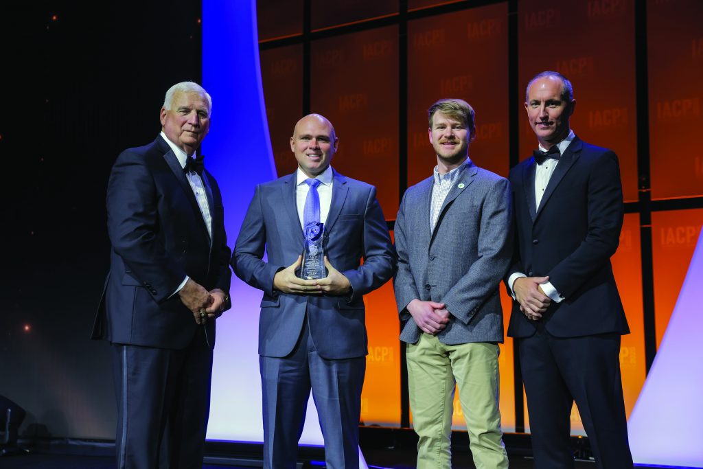 Four individuals in formal attire stand on stage with orange IACP backdrop; center person holds a glass award while others pose under bright event lighting.