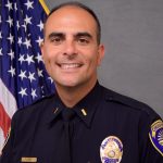 Uniformed officer stands before U.S. flag; visible badge, nameplate, and two pens in shirt pocket