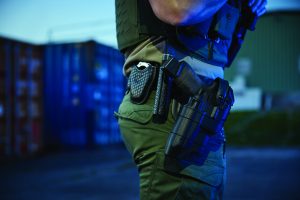 Close-up of a duty belt with holstered equipment worn over tactical pants, shown beside stacked shipping containers under blue lighting in an outdoor setting