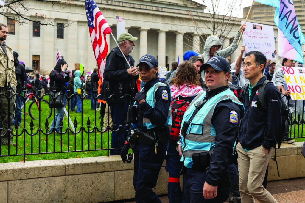 Columbus police dialogue team officers in reflective vests walk alongside protesters carrying signs and flags near a fenced lawn and columned government building