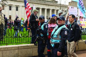 Columbus police dialogue team officers in reflective vests walk alongside protesters carrying signs and flags near a fenced lawn and columned government building