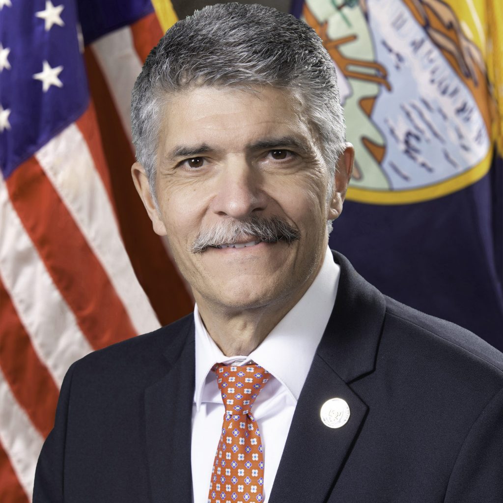 Man in dark suit, white shirt, orange tie stands before U.S. flag and emblem flag; formal setting suggests official role