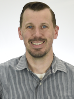 Studio portrait of an individual wearing a striped button‑down shirt over a light undershirt, shown from chest up against a plain white background with even lighting