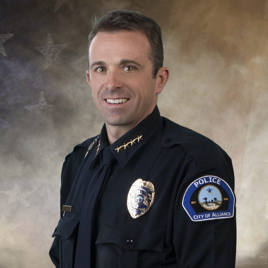 Police officer in dark uniform with gold insignia, badge, tie, and “City of Alliance” patch; faded American flag background
