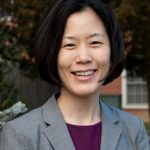 Person wearing a light gray blazer over a dark purple top, standing outdoors with trees and a brick building in the background